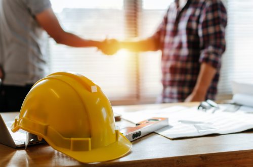 yellow safety helmet on workplace desk with construction worker team hands shaking greeting start up plan new project contract in office center at construction site, partnership and contractor concept